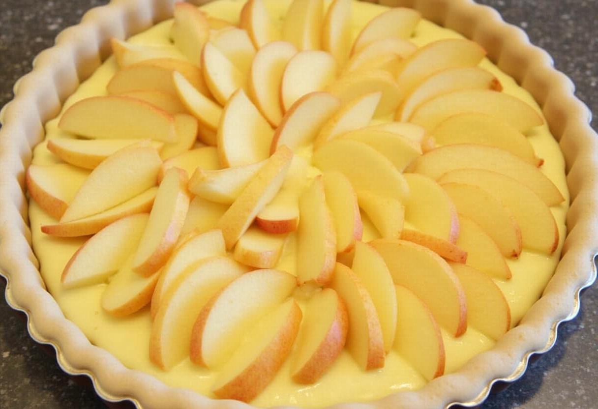 Assemblage de la tarte aux pommes crème avec une crème pâtissière étalée sur la croûte et les pommes disposées avec soin.